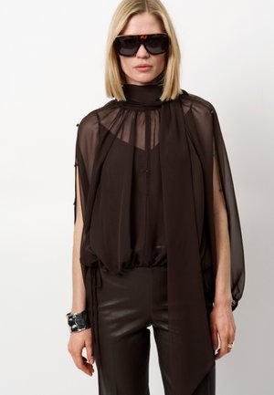 Zwarte doorschijnende top met hoge hals en gedrapeerde mouwen, gecombineerd met strakke nep leren broek. Een zilveren manchette-armband voegt detail toe.