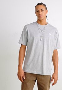 T-shirt gris à manches courtes en coton, avec un petit logo blanc sur la poitrine, associé à un pantalon marron. Design simple et décontracté.