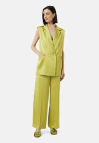 Limettengrünes satiniertes, doppelt geknöpftes Weste mit übergroßen Armausschnitten und Taschen, kombiniert mit weiten Hosen und grünen gerafften Sandalen.