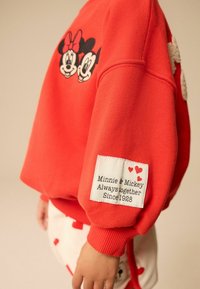 Punainen collegepaita, jossa on kirjailtu Minnie- ja Mikki Hiiren kasvot. Hihaan ommeltu etiketti sanoo: "Minnie & Mikki Aina yhdessä vuodesta 1928."