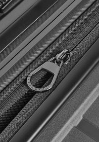 Détail de fermeture éclair de valise noire avec un tirette métallique marqué "EXPANDABLE" sur un fond de tissu texturé gris foncé.