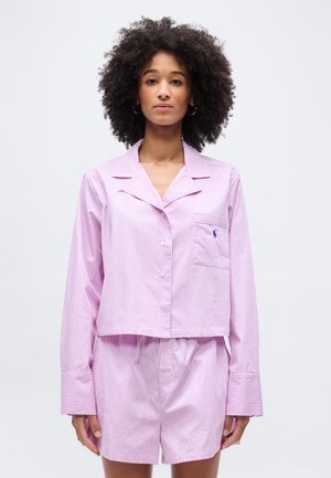 Vrouw met krullend haar, gekleed in een lichtroze en wit gestreept pyjamashirt met knopen en bijpassende korte broek, tegen een effen achtergrond.