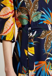 Klädsel med blommönster som har klara gula, gröna och blå blad på en marinblå bakgrund. Materialet verkar mjukt, med en knytning i midjan.