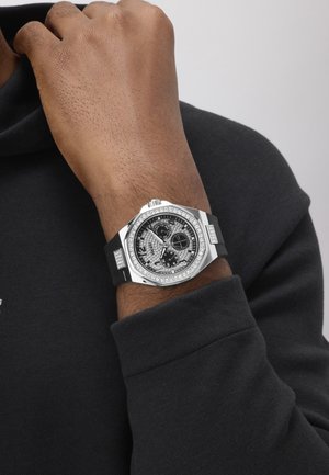 Wrist with black strap silver watch featuring a round diamond-studded bezel and three subdials on a black dial, worn over a black long sleeve.