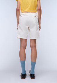 Weiße Baumwollshorts mit einem elastischen Taillenbund und seitlichen Taschen, mit einer Gesässtasche, die ein Logo trägt. Kombiniert mit blauen Socken und Schuhen.