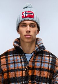 Cappello di lana azzurro chiaro con una toppa a forma di bandiera norvegese rossa, bianca e nera e pon-pon in cima, abbinato a una giacca in fleece a scacchi arancione e nera.