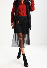 Femme portant un blouse rouge, un gilet en fausse fourrure noire, une jupe noire transparente avec des broderies florales, et des bottes ouvertes en chaussettes noires marchant en avant.