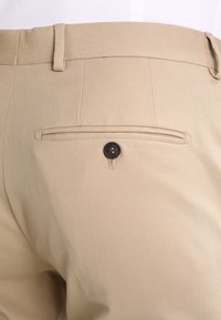 Beige skräddarsydda byxor med en slät yta, som har en bakficka med en svart knapp och detaljerad sömnad.