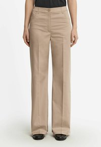Beige weite Hose aus glattem Material mit flachem Bund, zwei Seitentaschen und Knopfverschluss. Getragen mit schwarzem Schuhwerk.