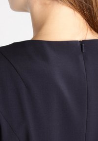 Robe bleu marine avec un col montant, une texture lisse et une fermeture à glissière invisible dans le dos, présentant des détails discrets de coutures.