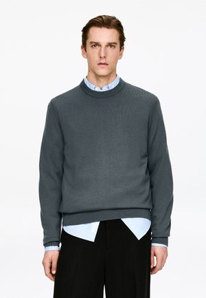 Dark grey knit sweater with a round neckline. Worn over a light blue collared shirt. Features long sleeves and a relaxed fit.