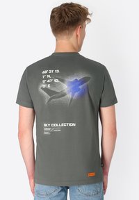 Grijze katoenen T-shirt met een grafisch ontwerp van een vliegtuig en coördinaten op de achterkant. Heeft een relaxte pasvorm en een klein oranje merktag.