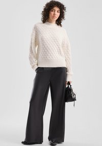 Beiger Strickpullover mit hohem Kragen, kombiniert mit schwarzen, weit geschnittenen Hosen. Das Model hält eine kleine schwarze Handtasche.