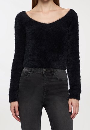 Femme portant un pull noir duveteux à manches longues et épaules dénudées, ainsi qu'un jean gris foncé taille haute, debout avec les mains détendues.