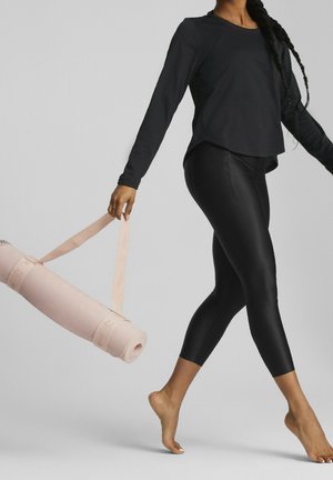 Osoba idąca boso, trzymająca zrolowaną różową matę do jogi z paskiem, ubrana w czarną bluzkę z długim rękawem i czarne legginsy.