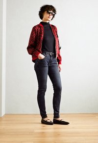 Červená a černá kostkovaná bomber bunda přes černý rolák, doplněná tmavými džíny a černými balerínami v minimalistickém prostředí.