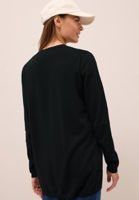 Zwarte longsleeve top met een ronde hals en een ontspannen pasvorm. Iets langere achterzoom, gladde textuur en zachte stof. Gemaakt met een pet.