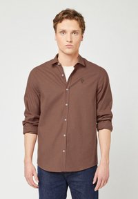 Camicia marrone a maniche lunghe con un motivo a quadretti discreto, in tessuto leggero, e un piccolo logo ad alberello sul petto. Abbinata a jeans scuri.