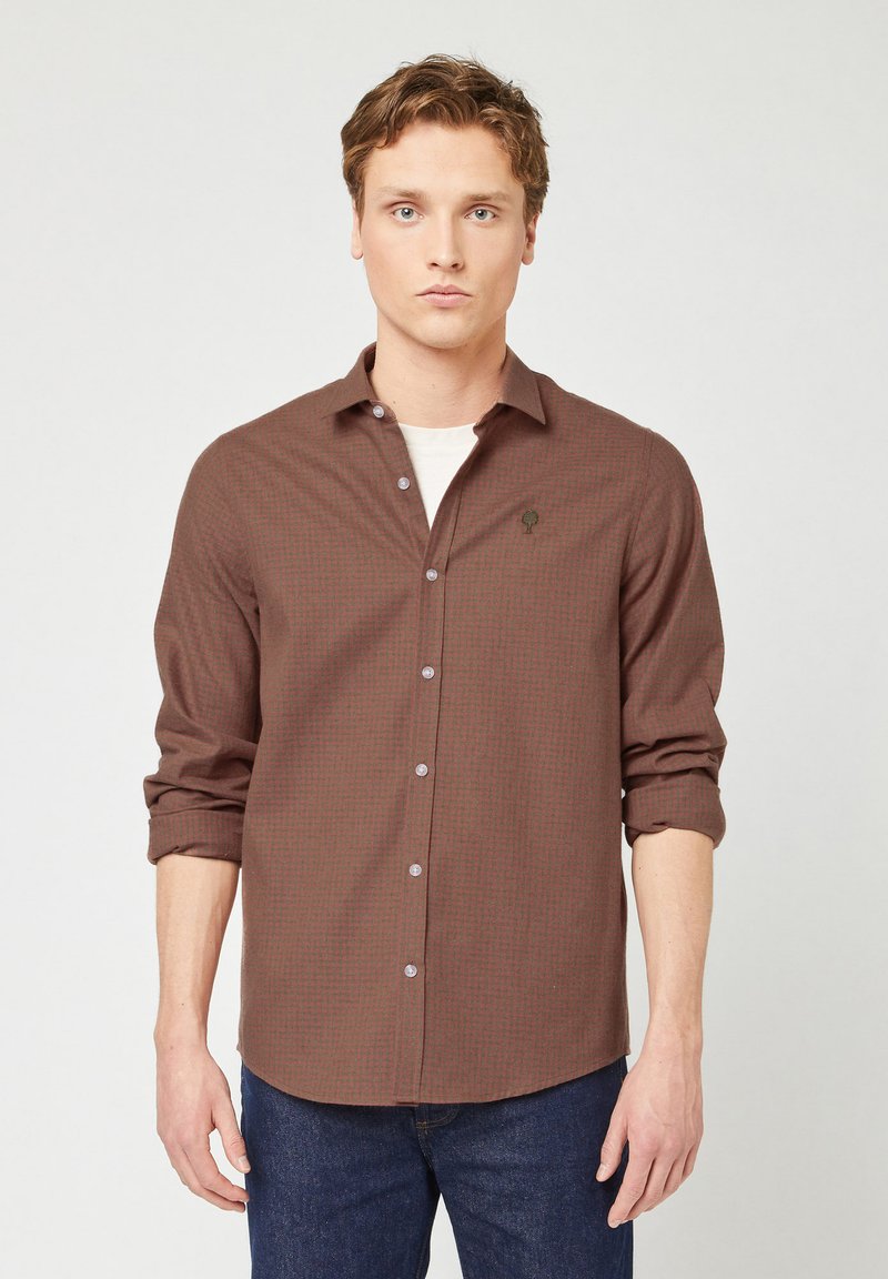 Camicia marrone a maniche lunghe con un motivo a quadretti discreto, in tessuto leggero, e un piccolo logo ad alberello sul petto. Abbinata a jeans scuri.