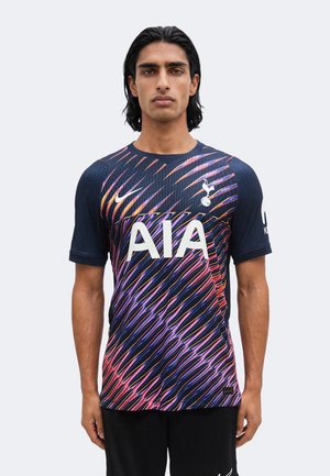 Mężczyzna stojący przodem, ubrany w ciemnoniebieską koszulkę piłkarską Tottenham Hotspur z wielobarwnymi ukośnymi paskami i białymi logo sponsorów.