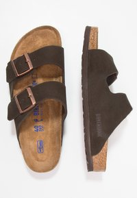 Sandali in pelle marrone con due cinturini regolabili, fondello in sughero e suola in gomma texturizzata. Presentano un logo impresso e una finitura in camoscio.