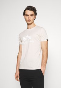 Světle béžové tričko z měkké bavlny, s velkým bílým logem a nápisem "Alpha Industries" na hrudi. Krátké rukávy a kulatý výstřih.
