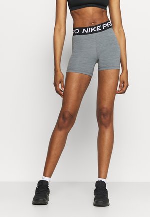 Atletisch persoon die grijze Nike Pro shorts, een zwarte sportbeha, witte sokken en zwarte sportschoenen draagt, staand op een eenvoudige witte achtergrond.