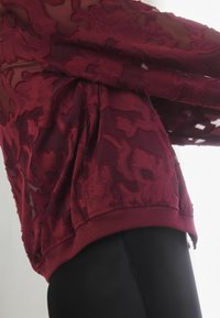 Haut bordeaux avec une superposition en dentelle florale, présentant une ceinture côtelée et un tissu texturé, partiellement superposé à un vêtement noir.