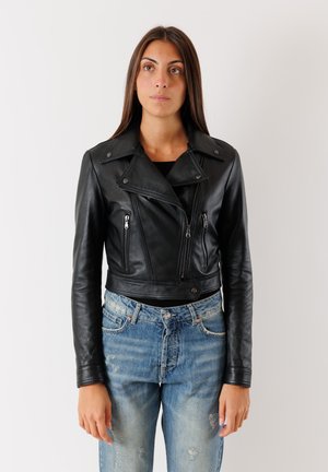 Donna con capelli lunghi e castani indossa una giacca nera da motociclista in pelle e jeans blu consumati, in piedi davanti a uno sfondo chiaro e uniforme.