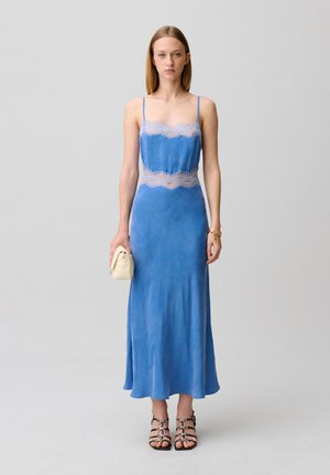 Giovane donna con un vestito a sottoveste blu con dettagli in pizzo, che tiene una piccola clutch beige, indossa sandali neri con cinturini e un braccialetto d'oro.