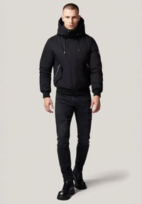 Czarna pikowana kurtka z odpinanym kapturem, zapinanymi kieszeniami, ściągaczami przy mankietach, noszona z przetartymi czarnymi dżinsami i masywnymi czarnymi butami.