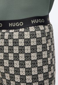 Κοντινό πλάνο μέσης που δείχνει ελαστική μέση με ετικέτα «HUGO» πάνω από καρό ύφασμα με επαναλαμβανόμενο κείμενο «HUGO» σε μαύρα και μπεζ τετράγωνα.