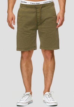 Shorts en coton vert olive avec une taille élastique, fermeture à cordon, et poches latérales, associés à des baskets blanches.