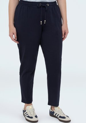 Person iført marineblå joggebukser med snor i taljen og beige og marineblå Adidas-sneakers på hvid baggrund.