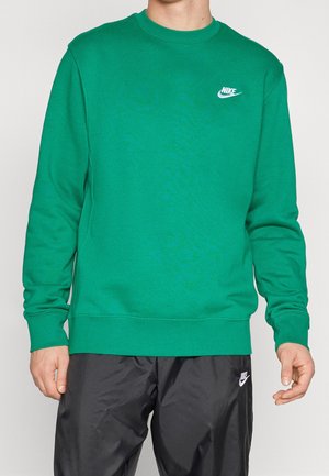 Mężczyzna w zielonej bluzie Nike i czarnych spodniach Nike, stojący z rękami luźno opartymi po bokach na jednolitym tle.