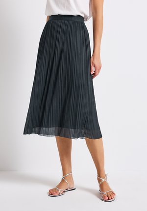 Jupe midi plissée en gris foncé, dotée d'une large taille et d'un ourlet fluide. Associée à des sandales à lanières argentées pour un look complet.