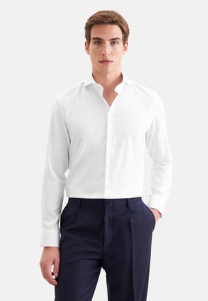 Mann trägt ein eng anliegendes weißes Hemd mit Knöpfen und dunkelblaue Hosen, steht mit einer Hand in der Tasche vor einem einfarbigen Hintergrund.