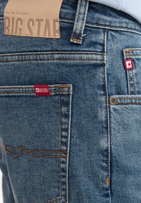 Vaqueros de mezclilla en azul medio con costuras naranjas en contraste. Presentan etiquetas de marca rojas y una etiqueta de cuero marrón en la cintura trasera.