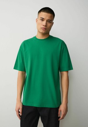 Noor mees seisab palja seina ääres, seljas lihtne roheline lühikeste varrukatega t-särk ja mustad püksid, vaatab ette neutralse ilmega.