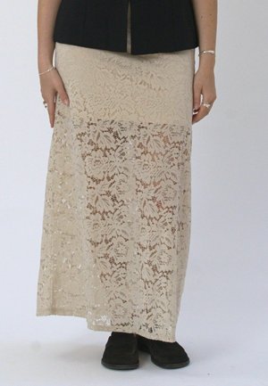 Top negro combinado con una falda beige de encaje floral y zapatos negros, manos relajadas a los lados con pulseras y anillos plateados.