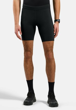 Homme portant un short de compression athlétique noir, des chaussettes noires montant jusqu'au mollet et des chaussures de course noires, debout devant un fond clair uni.