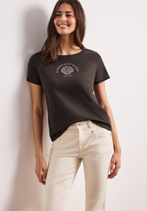 Femme souriante appuyée contre un mur, portant un t-shirt noir avec un motif coquillage et le texte « L'ÉTÉ EST MA SAISON » et un pantalon beige clair.