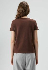 Braunes Baumwoll-T-Shirt mit kurzen Ärmeln, rundem Ausschnitt und legerer Passform, kombiniert mit hellgrauer Hose. Glatte Textur und schlichtes Design.