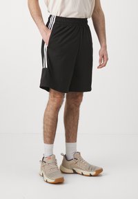 Man i svarta träningsshorts med vita ränder, vita strumpor och beige sneakers som står mot en enfärgad bakgrund.
