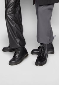 Dos personas que llevan botas negras con cordones y suelas gruesas; una lleva pantalones de cuero negros, la otra lleva pantalones de tela gris con puños elásticos.