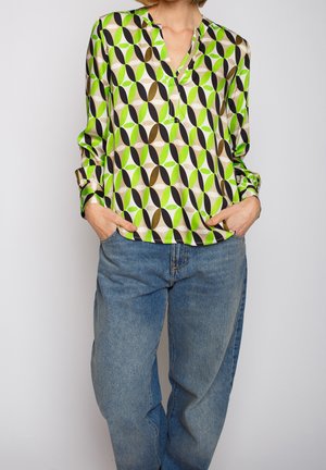 Persona che indossa una camicetta a maniche lunghe con motivi verdi, neri e beige e jeans blu larghi con le mani nelle tasche.