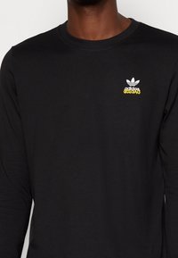 Svart långärmad skjorta i mjukt tyg med en vit tre-stripes logotyp och färgglad "adidas Originals" text på bröstet.