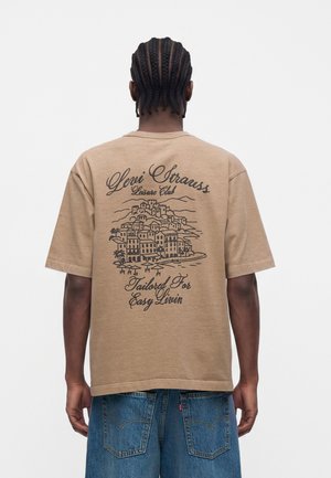 Άνδρας με πλεξούδες φοράει μπεζ μπλουζάκι Levi Strauss με εικόνα πόλης και κείμενο "Leisure Club, Tailored For Easy Livin" στο πίσω μέρος.