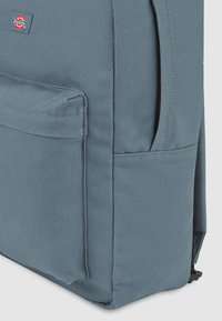 Mochila de tela azul con una superficie texturizada, bolsillo lateral y parche con logo. Presenta una forma cuadrada y costuras resistentes para mayor durabilidad.
