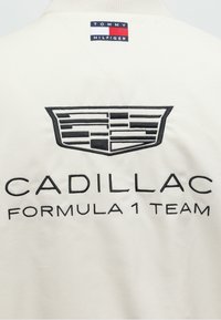 Λευκό ύφασμα με μαύρο κεντημένο λογότυπο Cadillac και κείμενο "Cadillac Formula 1 Team,", καθώς και μια μικρή ετικέτα Tommy Hilfiger κοντά στον γιακά.